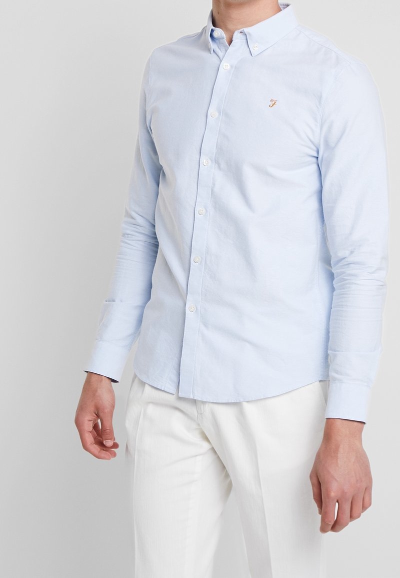 Camisa de manga larga azul claro con cuello clásico, que presenta un pequeño logo en el pecho y botones en la parte delantera.