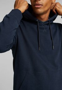 Sudadera con capucha en navy, con bolsillo frontal, capucha con cordón y tejido texturizado. Presenta costuras reforzadas y extremos de metal en el cordón.
