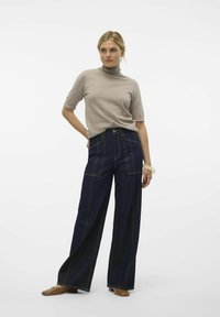 Bež turtleneck pulover v kombinaciji z visokimi širokimi temno modrimi kavbojkami, ki imajo kontrastno šivanje in sprednje žepke. Rjave čevlje.