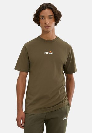 Jugendlicher mit lockigem Haar trägt ein olivgrünes Ellesse-T-Shirt und passende Hosen und steht vor einfachem Hintergrund.