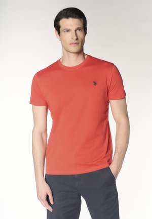 Uomo che indossa una maglietta rossa semplice a maniche corte con un piccolo logo scuro sul petto e pantaloni scuri, in piedi con una mano in tasca.