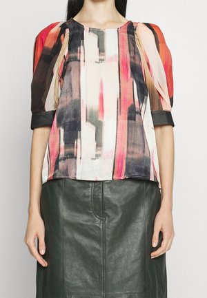 Femme portant un chemisier à motif abstrait coloré avec des manches bouffantes et une jupe en cuir gris foncé, debout devant un fond uni.