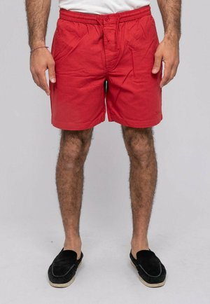 Homme portant un short rouge décontracté, un t-shirt blanc et des chaussures noires sans lacets, debout les mains relâchées le long du corps sur un fond gris uni.