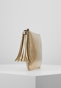 Kultainen metallinhohtoinen pussukka, jossa on sile ä pinta, vetoketjusuljin ja koristeelliset hapsut sivulla.