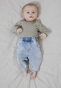 Camisa de rayas de manga larga en verde oliva y blanco, combinada con pantalones de mezclilla azul claro. Bebé tumbado sobre una superficie gris clara.