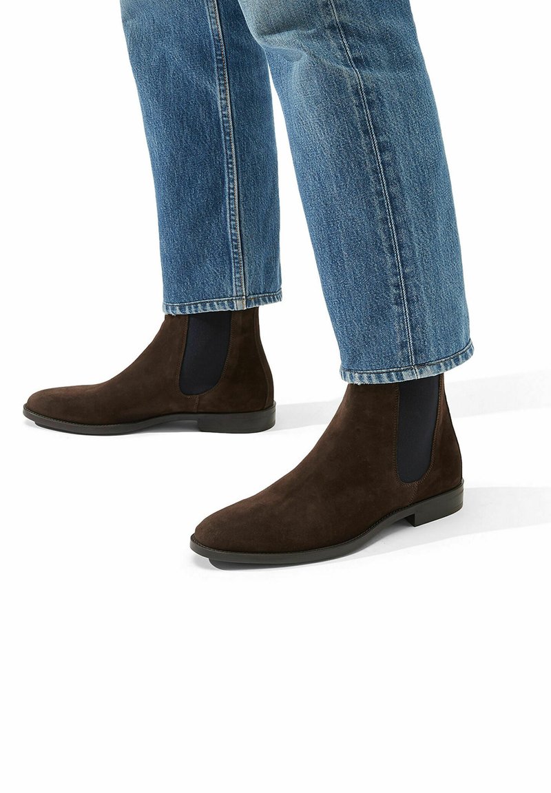 Botas Chelsea de ante marrón con paneles laterales elásticos, suelas planas y una silueta elegante, combinadas con unos jeans de tiro recto y puños en color azul claro.