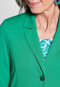 Grüner Blazer mit strukturiertem Design, Revers und einem einzelnen Knopfverschluss. Darunter befindet sich ein gemustertes Oberteil mit türkisfarbenen und weißen Akzenten.