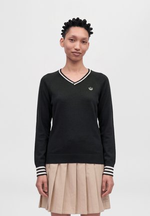 Un modèle porte un pull noir à col en V, orné de bordures rayées et d’un petit logo sur la poitrine, avec une jupe plissée beige, sur un fond uni.