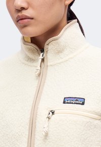 Κοντινό πλάνο ενός ατόμου που φορά μια μπεζ fleece μπουφάν Patagonia με φερμουάρ στην τσέπη στο στήθος και ψηλό γιακά.