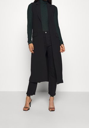 Donna che indossa un lungo cappotto nero senza maniche, dolcevita verde scuro, pantaloni neri e sandali neri con tacchi alti in piedi su uno sfondo bianco.