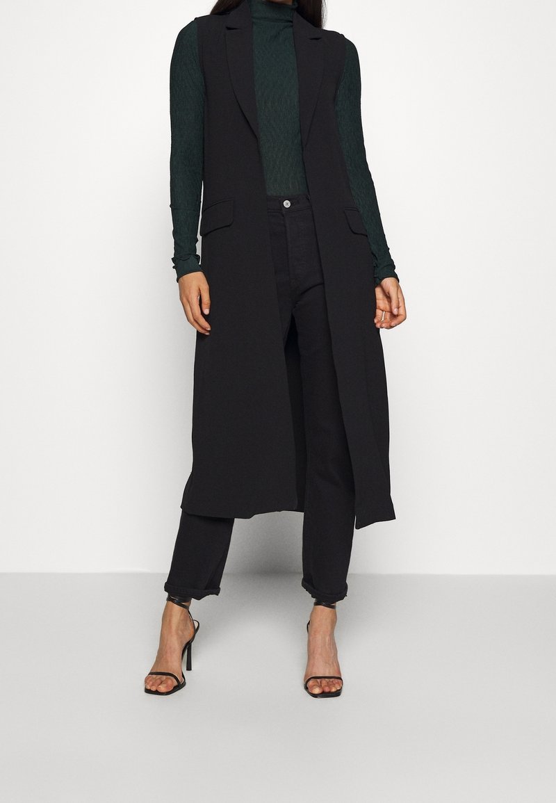 Femme portant un long manteau noir sans manches, un col roulé vert foncé, un pantalon noir et des sandales noires à talons hauts, debout sur fond blanc.