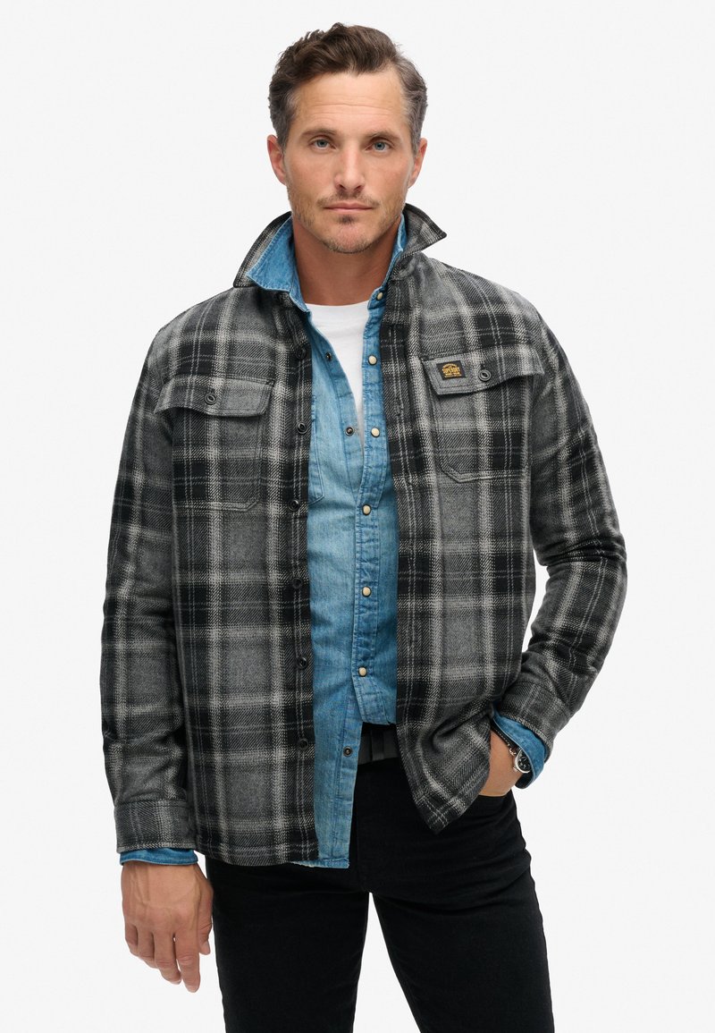 Chemise en flanelle à carreaux noirs et gris avec deux poches sur la poitrine portée sur une chemise en jean bleu clair boutonnée. Pantalons en jean noir et col texturé.