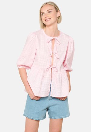 Ružová kockovaná blúzka s krátkymi nafúknutými rukávmi, viazaným výstrihom a siluetou peplum, v kombinácii so svetlými džínsovými šortkami.