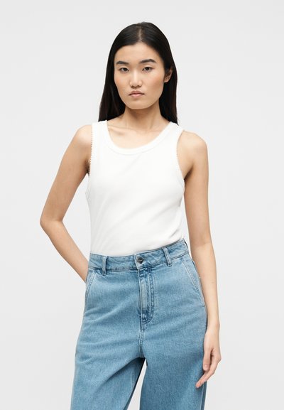 Νέα γυναίκα με μακριά σκούρα μαλλιά, ντυμένη με λευκό αμάνικο τοπ και ψηλόμεσα φωτεινά μπλε τζιν, στέκεται μπροστά σε ένα απλό φόντο.