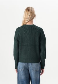 Lys hår person som bærer en mørkegrønn ribbestrikket genser og blå jeans sett fra baksiden mot en enkel hvit bakgrunn.