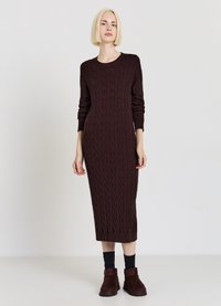 Robe en tricot bordeaux avec un motif torsadé ; manches longues, encolure ronde, coupe ajustée, longueur mi-mollet. Portée avec des chaussettes foncées et des bottes.