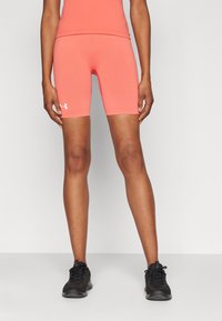 Osoba v koralových priliehavých šortkách Under Armour a zodpovedajúcom topu, stojaca s rozkročenými nohami, v čiernych športových topánkach, na bielom pozadí.