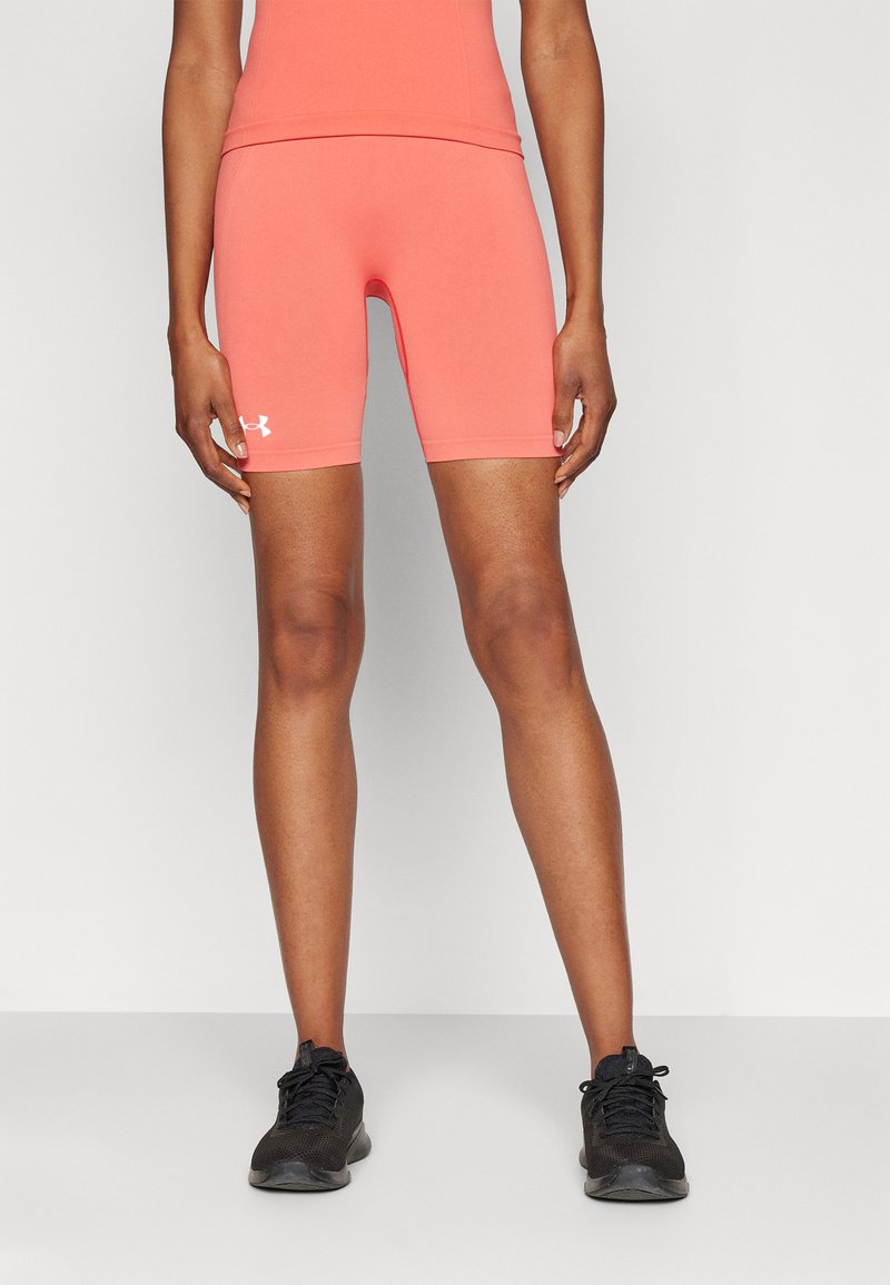 Osoba v koralových priliehavých šortkách Under Armour a zodpovedajúcom topu, stojaca s rozkročenými nohami, v čiernych športových topánkach, na bielom pozadí.