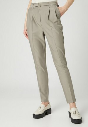 Schmal geschnittene beige Hose mit vorderen Abnähern und Gürtelschlaufen, getragen mit weißen Tassel-Loafers vor hellem Hintergrund.