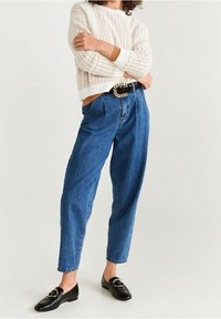 Cremfarbener Pullover mit Struktur, hochgeschnittene blaue Denim-Jeans in lässiger Passform, schwarzer Gürtel mit goldener Kettendetail und schwarze Slipper.