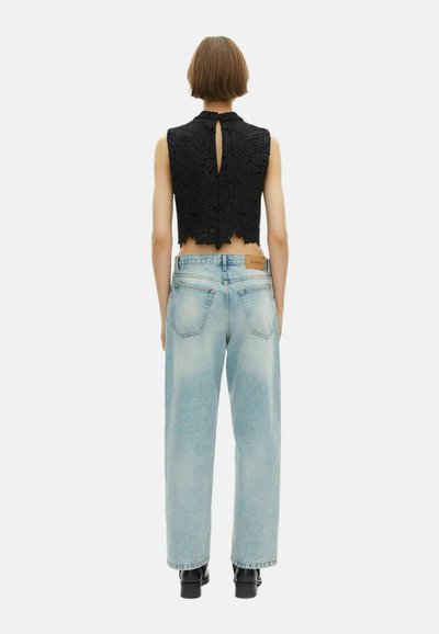 Person mit kurzen Haaren, die ein ärmelloses schwarzes Spitzen-Crop-Top und eine hellblaue High-Waist-weit geschnittene Jeans trägt und mit dem Rücken zu sehen ist, vor einem weißen Hintergrund steht.