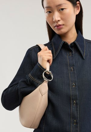 Nainen, jolla on päällään tumma denimpaita, kantaa olkapäällä beigeä nahkalaukkuja, joissa on hopeinen rengasdetaili, neutraalia taustaa vasten.