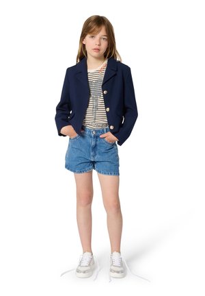 Jong meisje staand met handen in de zakken, gekleed in een marineblauw colbert, een gestreept T-shirt met olifantafbeelding, spijkershorts en witte sneakers met losgeknoopte veters.