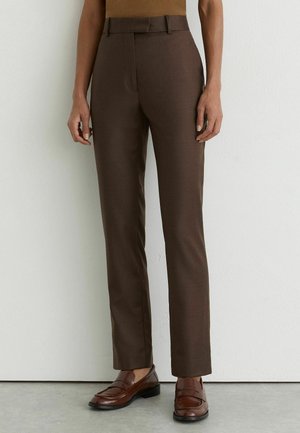 Straight-cut brown trousers worn with brown leather loafers on a person standing against a white wall.