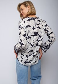 Bluse mit langen Ärmeln in einem floralem Muster aus Creme und Marineblau, mit gemusterten Manschetten und rundem Ausschnitt. Getragen mit hellblauen Jeans.