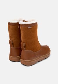 Bottines en daim marron avec une douce doublure en fourrure blanche, une semelle en caoutchouc et une petite étiquette portant la mention "Camper" près du haut.