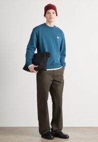 Felpa turchese con polsini a coste e un piccolo logo, abbinata a pantaloni verde oliva. Il modello tiene in mano una borsa di stoffa nera, indossando un cappello borgogna.