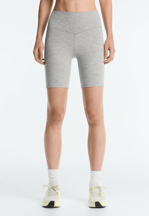 Kvinde iført lysegrå tætsiddende atletikshorts til midt på låret, hvide sokker og hvide løbesko, stående med hænderne afslappet ned langs siderne.