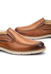 Zapatos de cuero marrón tipo mocasín con punta redondeada, suela de goma beige y costuras decorativas a los lados. Textura suave con acentos mínimos.