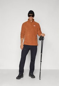 Pulôver bege de lã com fecho quarter zip, combinado com calças pretas e botas de caminhada. A figura segura um bastão de trekking.