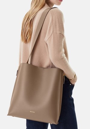 Mujer con suéter de cuello alto beige y jeans oscuros lleva un gran bolso de cuero taupe con cremallera dorada y logo.