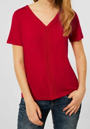 Femme portant un t-shirt rouge à col en V et manches courtes, un poing dans la poche et l'autre reposant près de sa cuisse.