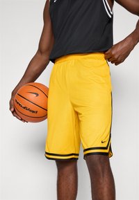 Gule basketball shorts lavet af meshstof, med sorte side-striber og en fleksibel talje. En basketball holdes i den ene hånd.