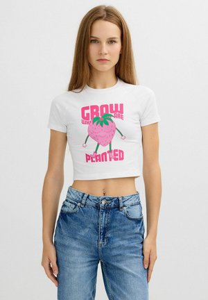 Mujer joven con top corto blanco con una fresa sonriente y el texto "Crece donde estás plantado", combinada con vaqueros azules de cintura alta.