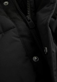 Detail čiernej izolačnej bundy s hladkým povrchom, viditeľným zipsom a cvokmi pozdĺž švu.