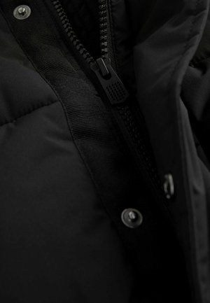 Svart isolerad jacka nära bild, med en slät textur, synlig dragkedja och tryckknappar längs sömmen.