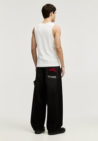 Débardeur blanc sans manches associé à un pantalon en denim noir oversized avec des accents graphiques rouges sur les poches arrière.