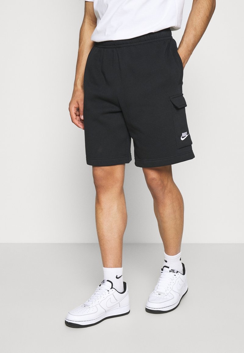 Svarta bomullsshorts med resår i livet, sidofickor och en cargoficka med en vit Nike-logga, tillsammans med vita sneakers.