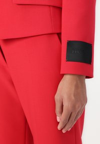Costume ajusté rouge avec une finition texturée, comportant une étiquette noire sur le poignet portant l'inscription "HUGO". Gros plan sur le détail de la manche et de la main.