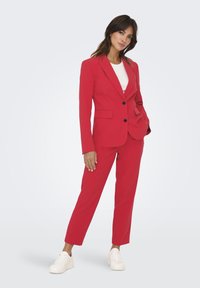 Costume rouge ajusté comprenant un blazer cintré avec deux boutons, des détails de poches et un pantalon droit, associé à des baskets blanches.