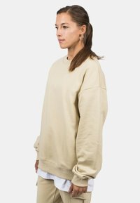 Felpa beige oversize con spalle scese e polsini a coste. Tessuto morbido con una texture liscia, indossata sopra una camicia bianca.