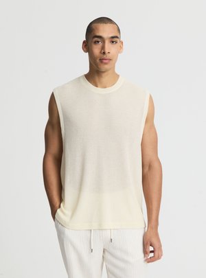 Giovane uomo con capelli corti che indossa un top lavorato a maglia crema senza maniche e pantaloni chiari, in piedi davanti a uno sfondo bianco semplice.
