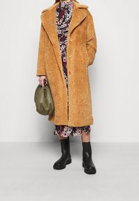 Ruskea tekoturkis takki, jossa on lovettu kaulus, nappikiinnitys ja sivutaskut, kerrostettuna kukkamekon päälle. Yhdistetty mustien nilkkureiden ja oliivinvihreän laukun kanssa.