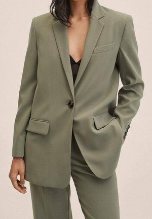 Femme portant un costume ajusté vert olive avec une veste à bouton unique et un pantalon assorti, une main dans la poche, top noir en dessous.