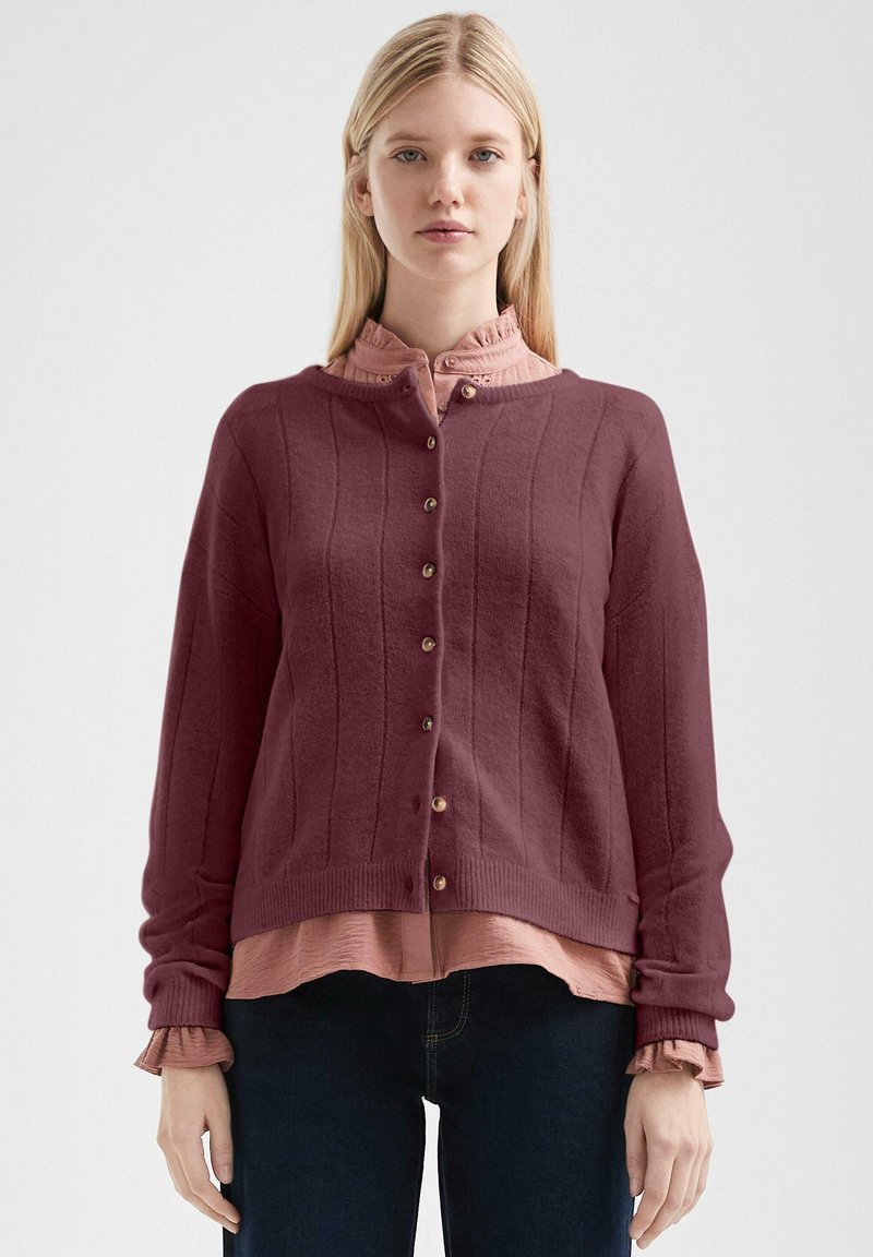 Bordeaux gebreide cardigan met geribbelde textuur, met een ronde kraag en een knoopsluiting aan de voorkant, gelaagd over een roze rucheblouse.