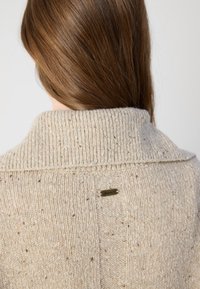 Sweater bege tricotado com um colarinho alto, apresentando um padrão texturizado e uma pequena etiqueta de metal na parte de trás. Aparência suave e salpicada.